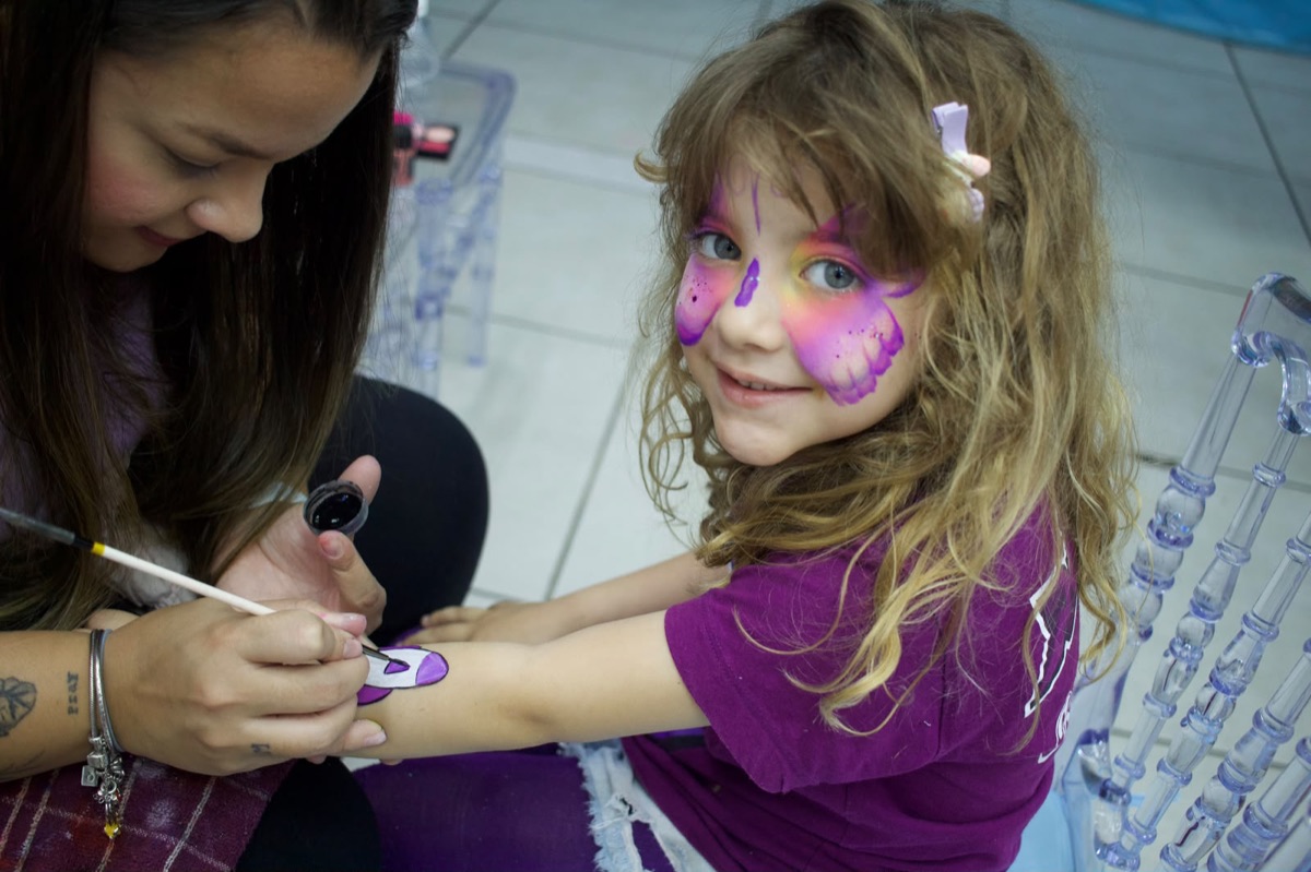Recreadora pintando o braço de uma menina que tem uma borboleta pintada no rosto